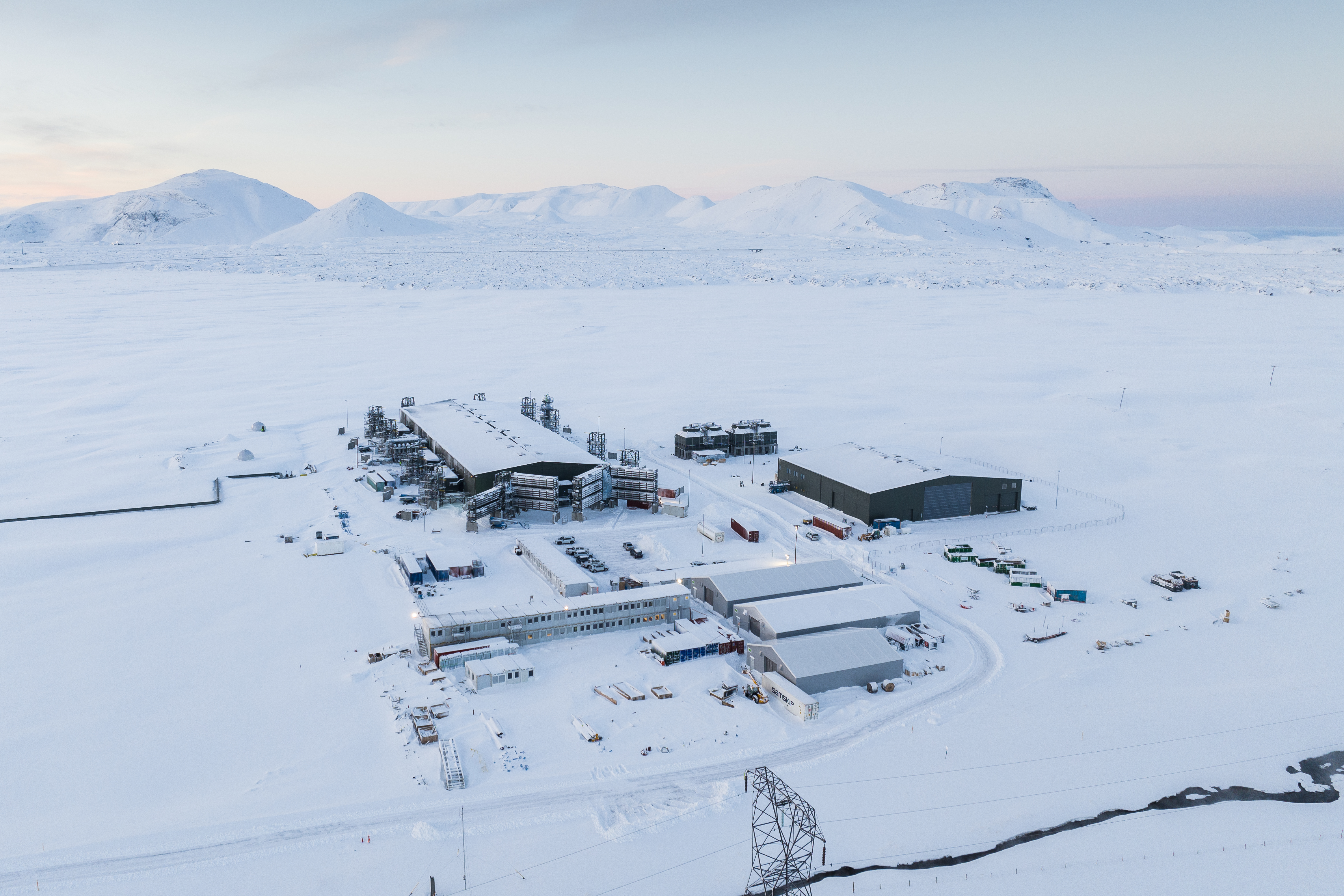 Oblique aerial photo of Climeworks' Mammoth direct air capture plant in Iceland.  Plant is comprised of a complex of buildings located on a flat snow-covered plain with distant mountains in background.