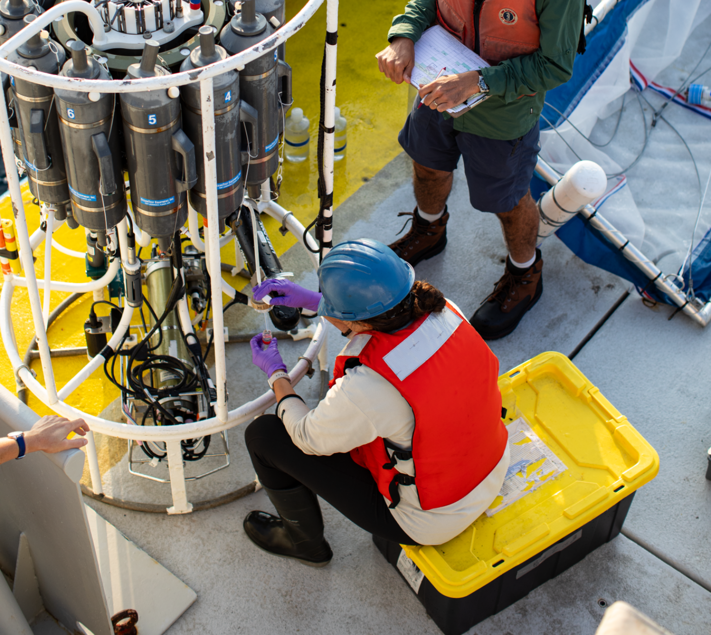 Photo looking down on two researchers working on the CTD device on the deck of the research vessel. The device is a bit larger than a person and is composed of several vertical metal tubes arranged in a circle, enclosed in a frame..
