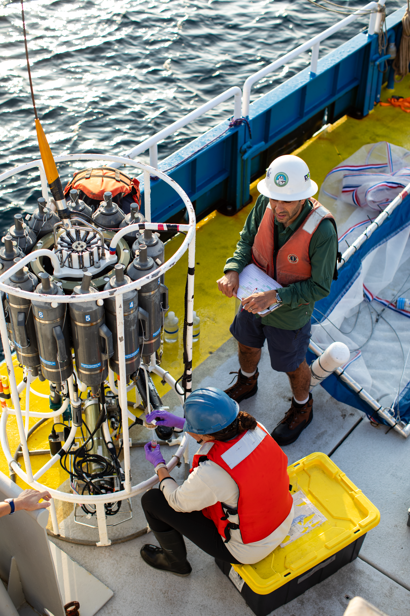 Photo looking down on two researchers working on the CTD device on the deck of the research vessel. The device is a bit larger than a person and is composed of several vertical metal tubes arranged in a circle, enclosed in a frame.